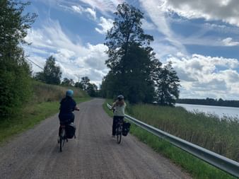 Zwei Radfahrer mit Packtaschen auf einer Schotterstraße zwischen Mariefred und Järna. Rechts sind ein großer Baum und ein See unter bewölktem Himmel sichtbar.