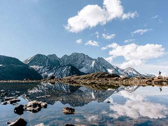 Eine Bergkulisse spiegelt sich in einem Bergsee. Im Hintergrund ein Wanderer.