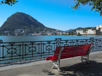 Banc en bois rouge sur la promenade du lac à Lugano avec balustrade en fer ornée, vue sur le lac turquoise et le Monte San Salvatore.