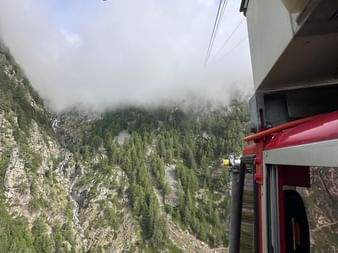 Blick aus einer rot-weißen Seilbahngondel über ein steiles bewaldetes Tal mit Felswänden. Nebel bedeckt die Berggipfel darüber.