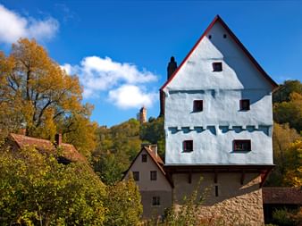 Weißes mehrstöckiges Mühlengebäude mit roter Dachkante im Taubertal, umgeben von Herbstbäumen und traditionellen Häusern unter blauem Himmel.