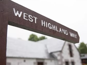 Brown wooden sign reading 'WEST HIGHLAND WAY' in white letters, mounted on wooden posts with blurred buildings in the background.