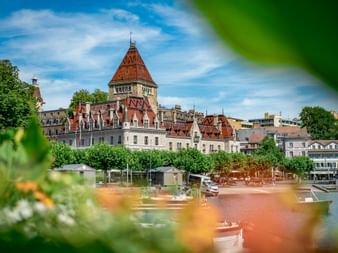 Château d'Ouchy avec toit de tuiles rouges et tour à Lausanne sur le Lac Léman, encadré par un feuillage vert et des fleurs colorées au premier plan.