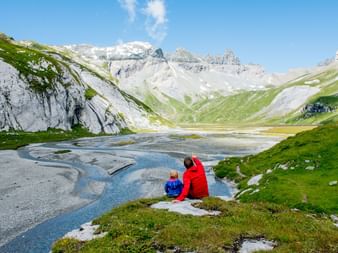 Mann und Kind sitzen auf einem Stein am Bach und blicken auf die Berglandschaft.