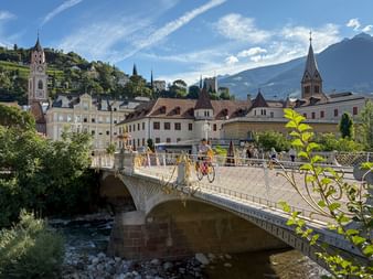 Radfahrer überqueren eine verzierte Steinbrücke über einen Fluss in Meran, mit historischen Gebäuden, Kirchtürmen und Bergen im Hintergrund unter blauem Himmel.