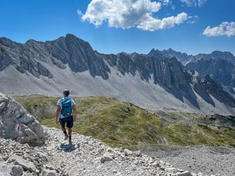 Wanderer mit Rucksack auf felsigem Bergpfad im Karwendel mit dramatischen Felsspitzen und Geröllhängen unter blauem Himmel mit Wolken.