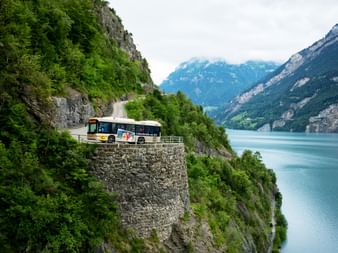 Bus on narrow stone-walled mountain road in Isenthal above turquoise Lake Lucerne, surrounded by green cliffs and distant mountains.