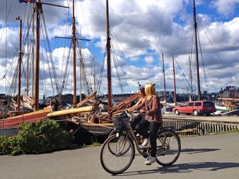 Zwei Radfahrer mit Fahrrädern stehen auf einer Uferpromenade in Stockholm, mit historischen Segelschiffen im Hafen hinter ihnen.
