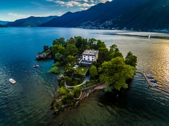 Luftaufnahme der Brissago Inseln im Lago Maggiore mit weißer Villa umgeben von üppiger grüner Vegetation, Boote in der Nähe, Berge im Hintergrund.