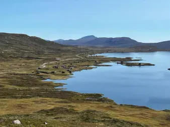 Ruhiger Bergsee mit kleinen Inseln im norwegischen Hochland. Sanfte Hügel und ferne Berge unter klarem Himmel entlang Mjølkevegen.
