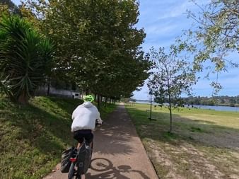 Radfahrer im weißen Hemd fährt auf baumgesäumtem Weg am Fluss zwischen Barcelos und Vila do Conde, mit grünem Flussufer und blauem Himmel.