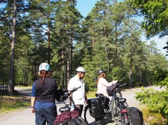 Drei Radfahrer mit Helmen und bepackten Rädern pausieren auf einem asphaltierten Weg durch einen Kiefernwald am Tag 3 der Stockholm-Rundtour.