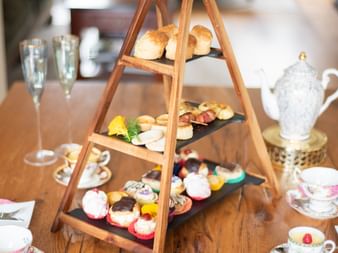 Hölzerner Etagenständer mit Scones, Gebäck und Kuchen auf einem Esstisch. Teetassen, Sektgläser und Beilagen vervollständigen das Afternoon-Tea-Setup.