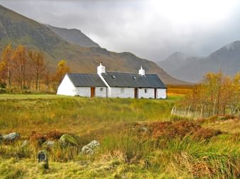 Weißes schottisches Cottage mit dunklem Dach im Highland-Tal, umgeben von Herbstbäumen und Bergen unter bewölktem Himmel nahe West Highland Way.