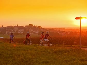 Quatre cyclistes avec casques se reposent sur une colline herbeuse au coucher du soleil doré, avec la ville historique de Morat en arrière-plan.
