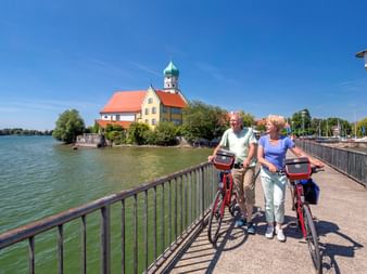 Deux cyclistes âgés avec vélos rouges sur promenade au bord du lac de Constance, château historique à dôme vert en arrière-plan.