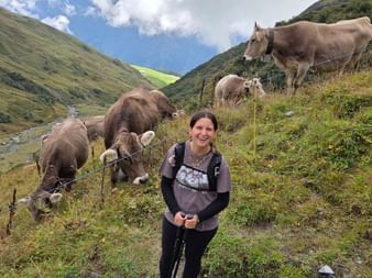 Wanderin Silvia lächelt in einem Alpental mit braunen Kühen auf der Weide. Grüne Berghänge und blauer Himmel mit Wolken im Hintergrund.