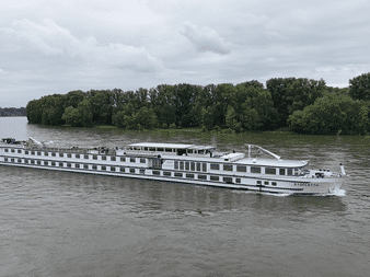 Navire de croisière fluviale MS Rigoletto blanc à plusieurs ponts naviguant sur une rivière aux eaux brunes avec une rive boisée verte.