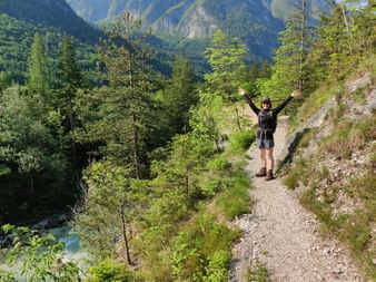 Eine Person in Wanderkleidung, Cap und Sonnenbrille streckt die arme aus. Sie steht auf einem Wanderweg umgeben von Bäumen, Bergen und einem Fluss.