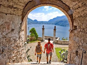 Zwei Touristen gehen durch Steinbogen in Morcote mit Blick auf Luganer See, Bergen und blauem Himmel im Hintergrund, Tessin, Schweiz.