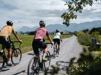 Three cyclists on gravel bikes riding along a paved path through green fields with mountains in the background in Gantrisch Nature Park.