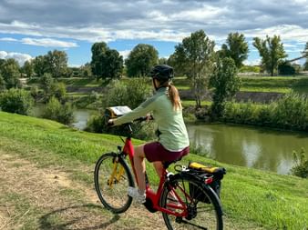 Cycliste avec casque et vélo rouge consultant une carte près d'un canal paisible entouré d'arbres verts sous un ciel nuageux.