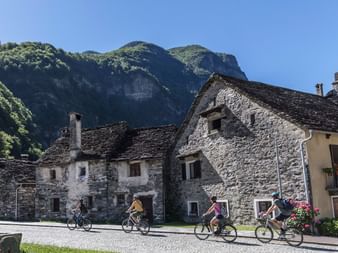 Gruppe von Radfahrern fährt durch traditionelles Steindorf in Vallemaggia, Tessin, mit historischen Gebäuden und grünen Bergen im Hintergrund.