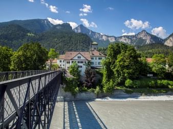 Brücke über den Rhein zwischen Reichenau-Bonaduz Metallbrücke über schlammigen Fluss mit Schweizer Alpendorf und dramatischen Berggipfeln im Hintergrund unter blauem Himmel mit weißen Wolken.