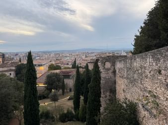 Alte Steinmauern und Zypressen mit Blick auf eine spanische Stadt in Nordkatalonien unter bewölktem Himmel.