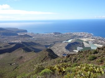 Vue panoramique de la côte de Tenerife depuis une position élevée montrant bâtiments blancs, océan bleu et paysage volcanique.