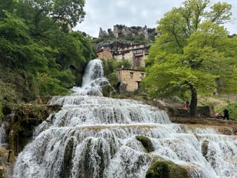 Kaskadierender Wasserfall fließt durch das Dorf Orbaneja del Castillo mit Steingebäuden und Felsklippen darüber, umgeben von grünen Bäumen.