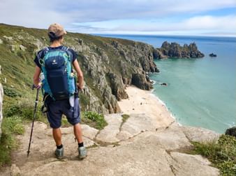 Randonneur avec sac à dos bleu et bâtons de marche sur falaise rocheuse surplombant mer turquoise et plage de sable en Cornouailles.