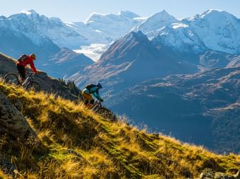 Deux coureurs de trail descendent la montagne sur un chemin naturel. En arrière-plan, un paysage de montagne enchanteur. Les sommets sont en partie recouverts de neige.