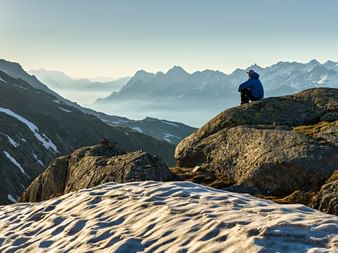 Une personne est assise sur des rochers et contemple le panorama des montagnes. Il y a encore de la neige sur les prairies de montagne