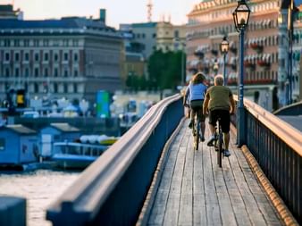 Zwei Radfahrer fahren über eine Holzbrücke in Stockholm während einer Radtour, mit historischen Gebäuden und Uferfront im Hintergrund.