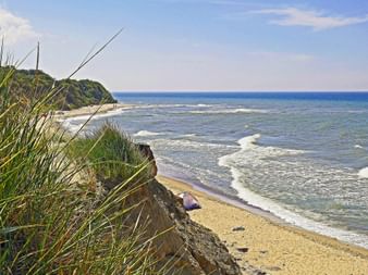 Vue sur la plage depuis le sentier de Rügen Vue côtière depuis une falaise de Rügen montrant plage de sable, mer Baltique bleue avec vagues blanches et côte boisée sous ciel nuageux.