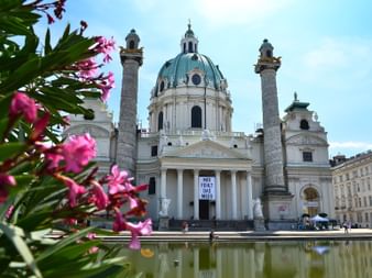 Karlskirche Barockkirche in Wien mit grüner Kupferkuppel, Zwillingssäulen und klassischem Portikus spiegelt sich im Wasserbecken. Rosa Blüten rahmen die Sicht.