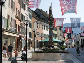Fußgängerzone in Delémont mit verziertem Brunnen, bunten Wappenflaggen, traditionellen Gebäuden mit Fensterläden und Passanten.