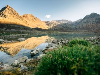 Ruhiger Bergsee in Graubünden spiegelt goldbeleuchtete Gipfel in der Morgensonne wider. Felsiges Ufer mit grüner Vegetation im Vordergrund.