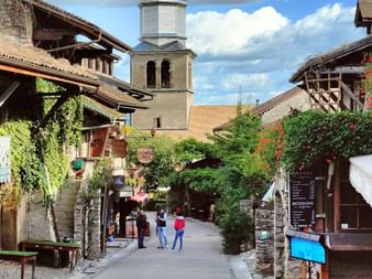 Kopfsteinpflasterstraße in Yvoire mit mittelalterlichen Steingebäuden, Geschäften und einem markanten Kirchturm mit Kegeldach unter bewölktem Himmel.
