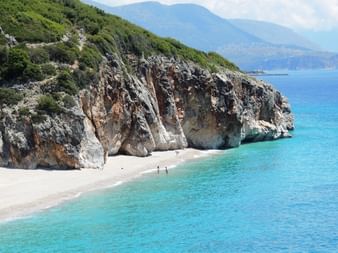 Weißer Kieselstrand unter dramatischen Felsklippen an der albanischen Küste. Türkisfarbenes Wasser und ferne Berge unter blauem Himmel.