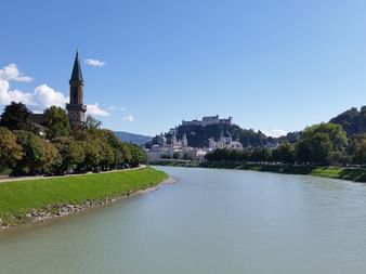 Blick entlang der Salzach in Österreich mit grünem Flussufer, Kirchturm und Bergfestung unter blauem Himmel mit weißen Wolken.