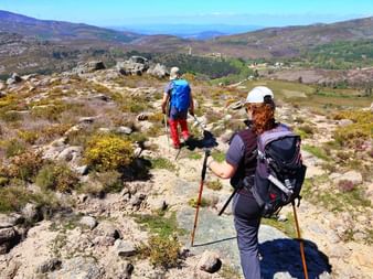 Zwei Wanderer mit Rucksäcken und Wanderstöcken auf felsigem Bergpfad im Peneda-Gerês-Nationalpark, Portugal, mit Hügeln und Tälern.