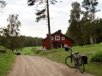 Tourenrad mit Taschen auf Schotterweg zu rotem Holzhaus umgeben von grünen Wiesen und Wald bei Stockholm.