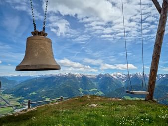 Bronzeglocke und Holzschaukel auf einem Berggipfel mit Alpenpanorama. Grüne Wiese mit Wildblumen, schneebedeckte Gipfel unter blauem Himmel.