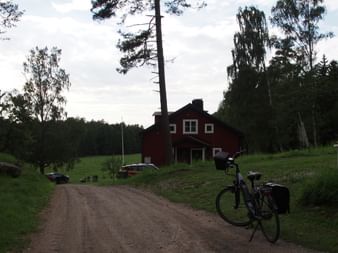 Tourenrad mit Packtaschen auf einem Schotterweg, der zu einem traditionellen roten Schwedenhaus führt, umgeben von grünen Wiesen und hohen Bäumen.