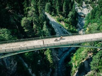 Aerial view of two cyclists on a metal bridge spanning a deep forested gorge in Graubünden, Switzerland, with a river flowing below.