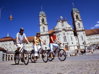 Drei Radfahrer mit Helmen auf Kopfsteinpflaster vor der barocken Abtei Einsiedeln mit Zwillingstürmen und ornamentaler weißer Fassade.
