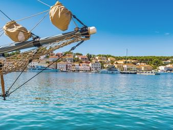 Blick vom Segelschiffbug mit Takelage und Netzen auf mediterrane Küstenstadt mit bunten Gebäuden und Booten im Hafen.