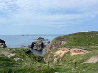 Zerklüftete Cornwall-Küste mit grünen, grasbewachsenen Klippen, Felsformationen im Meer und einem weißen Gebäude auf dem Hügel.